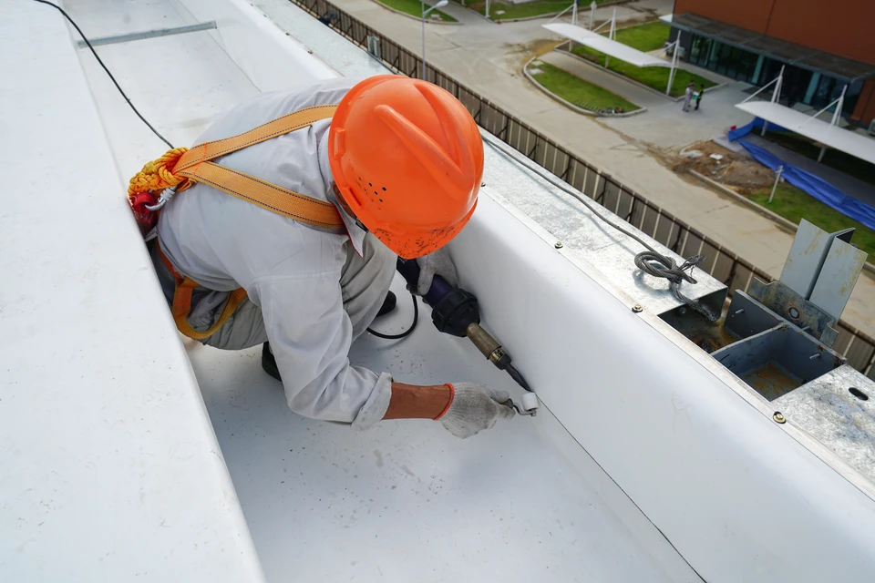 Ouvrier en combinaison blanche et casque orange travaillant sur toiture d'immeuble, équipé d'un harnais de sécurité
